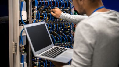 راهنمای انتخاب پیمانکار زیرساخت شبکه؛نمای پشت یک مرد جوان که در حال کار با ابررایانه است؛ کابل‌های سرور بلید را متصل می‌کند و همزمان داده‌ها را روی لپ‌تاپ بررسی می‌کند.